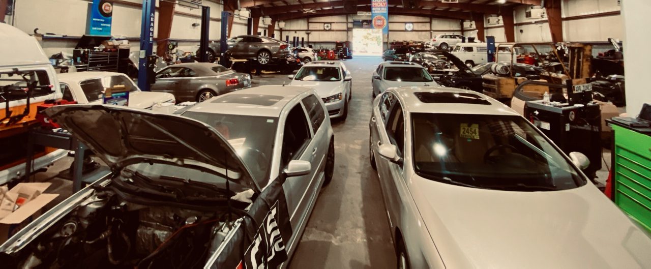 Auto repair shop interior with several cars, including a silver Volkswagen Golf and a white van.