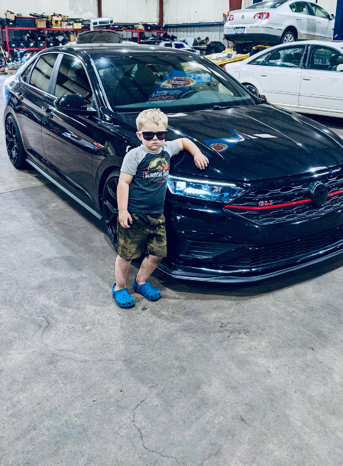 Young boy in sunglasses and Jurassic Park shirt leans on a black VW Jetta GLI.